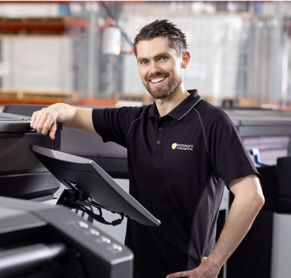 A man stands beside a large printer, showcasing its size and functionality in a professional setting.