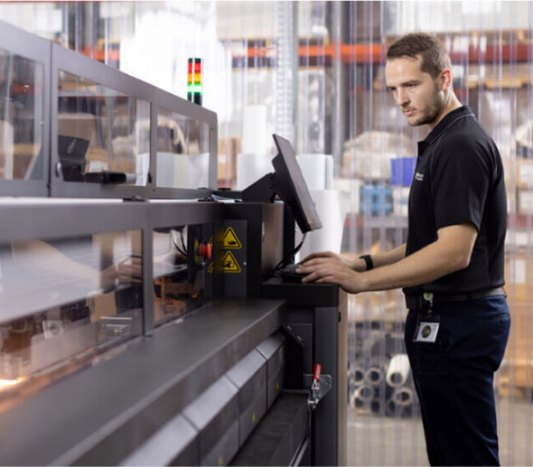 A man operates a large printer in a spacious warehouse, focused on his task amidst industrial surroundings.