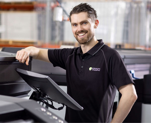 A man in a black shirt stands beside a printer, appearing focused and engaged in his task.