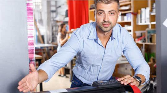 A man stands in front of a printer, observing the printing process with a focused expression.