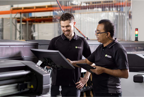 Two men in black shirts stand beside a large printer, engaged in conversation or collaboration.