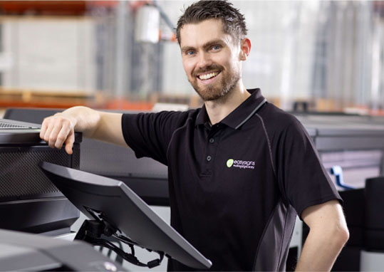 A man in a black shirt stands beside a printer, appearing focused and engaged in his task.