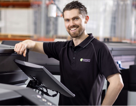 A man in a black shirt stands beside a printer, appearing focused and engaged in his task.