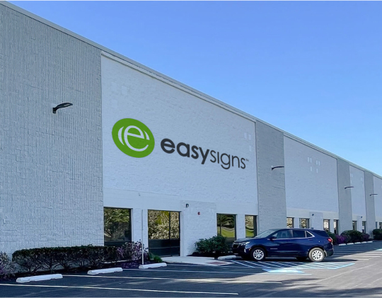 A view of the Easy Signs building located in a parking lot, showcasing its exterior and surrounding vehicles.