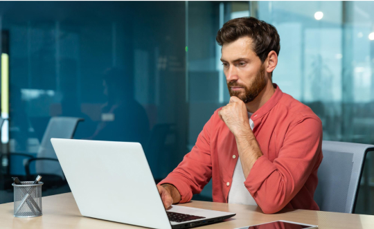 A man seated at a desk, focused on his laptop, surrounded by a tidy workspace and soft lighting.