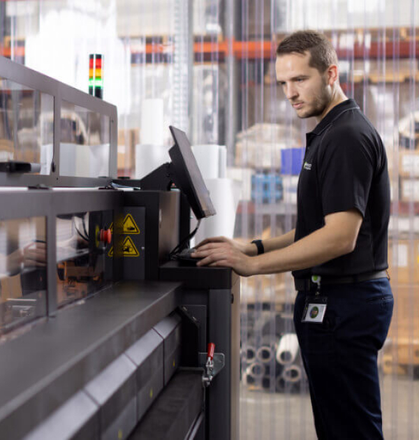 A man stands in front of a large printer, observing its operation with a focused expression.