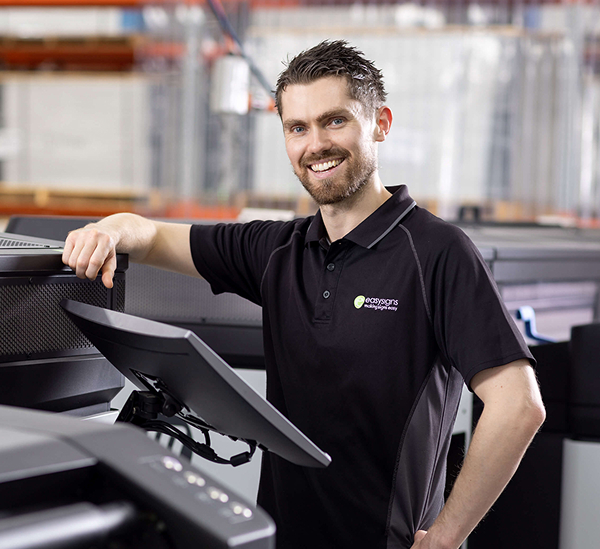 A man stands in front of a large printer, observing its operation with a focused expression.