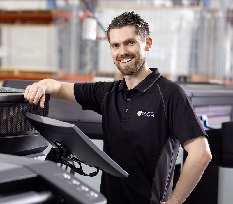 A man stands in front of a large printer, observing its operation with a focused expression.