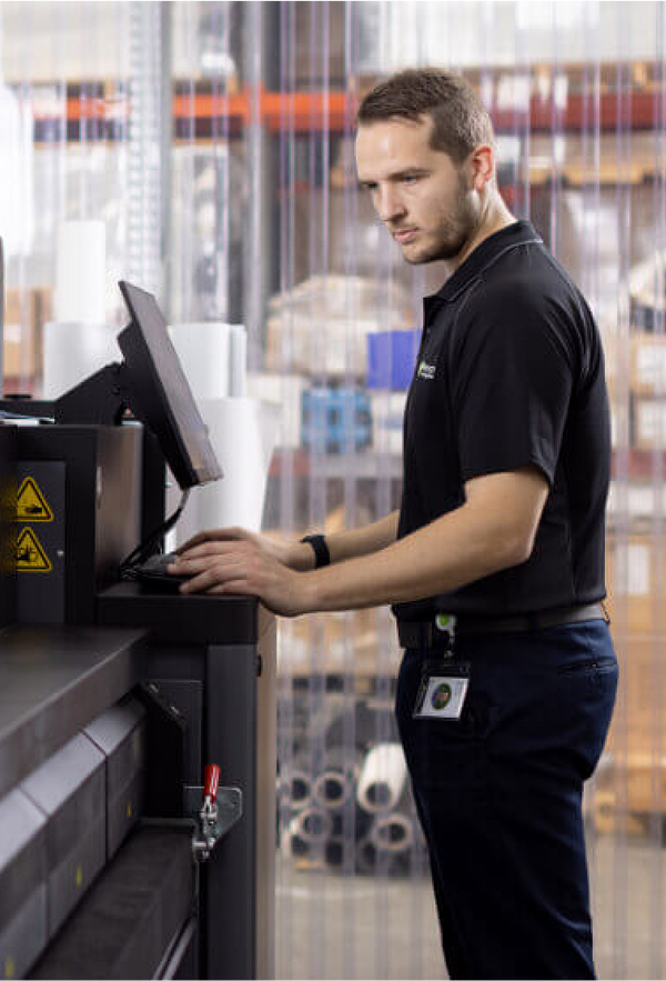 A man in a black shirt stands confidently in front of a large machine, showcasing a professional environment.