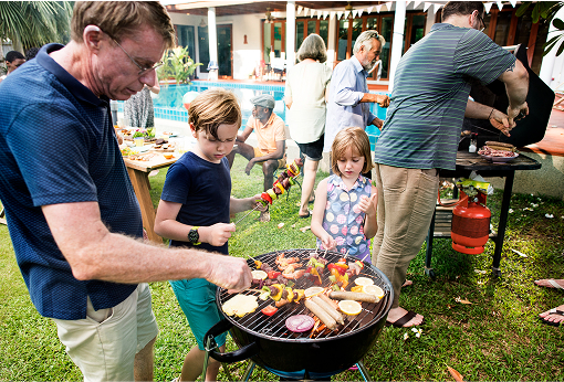 A man and two children joyfully cooking together on a barbecue, surrounded by a sunny outdoor setting.