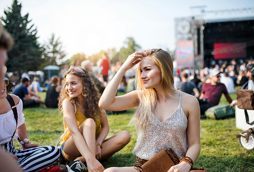 Three young women enjoying a music festival, seated on the grass, surrounded by vibrant festival atmosphere.