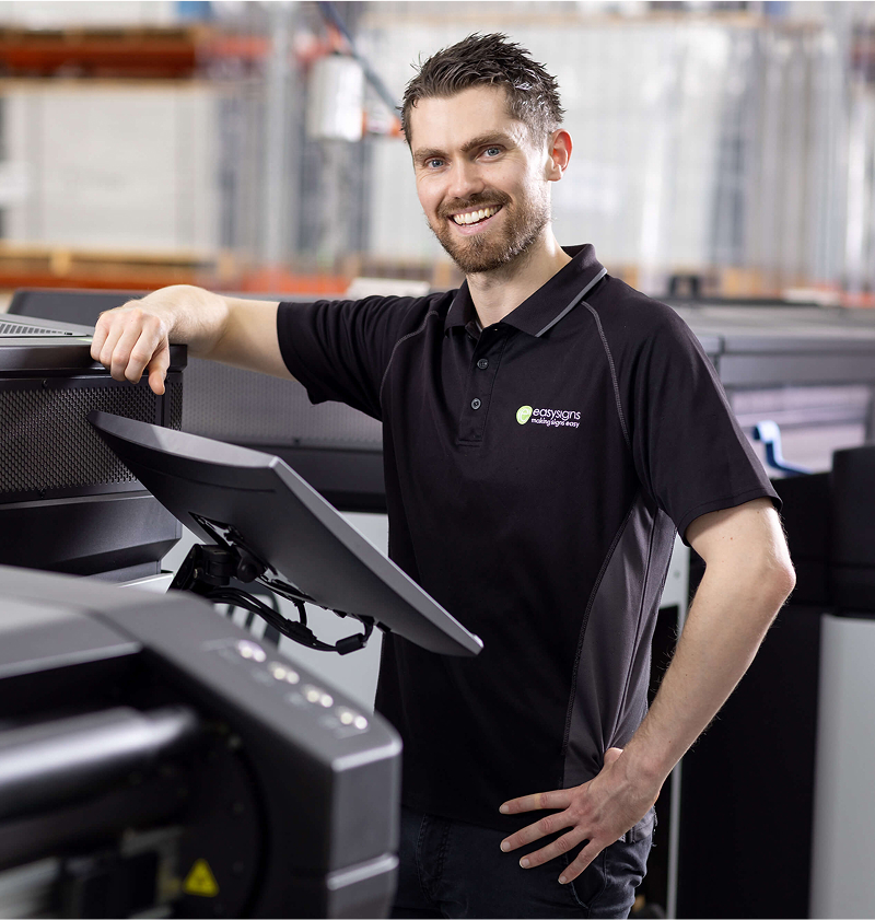 A man stands beside a large printer, showcasing the equipment in a professional setting.