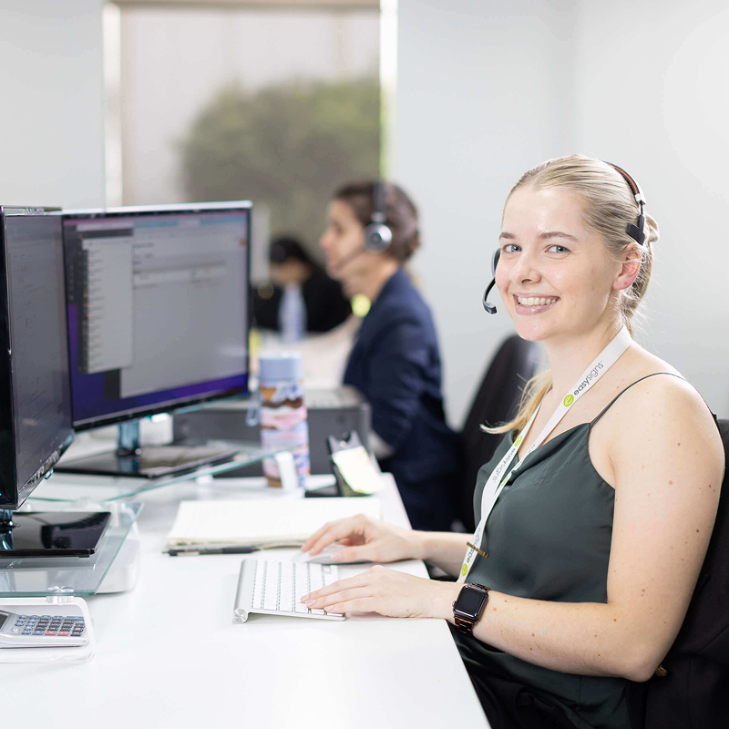 A woman wearing a headset sits at a desk, focused on her work, with a computer and office supplies around her.