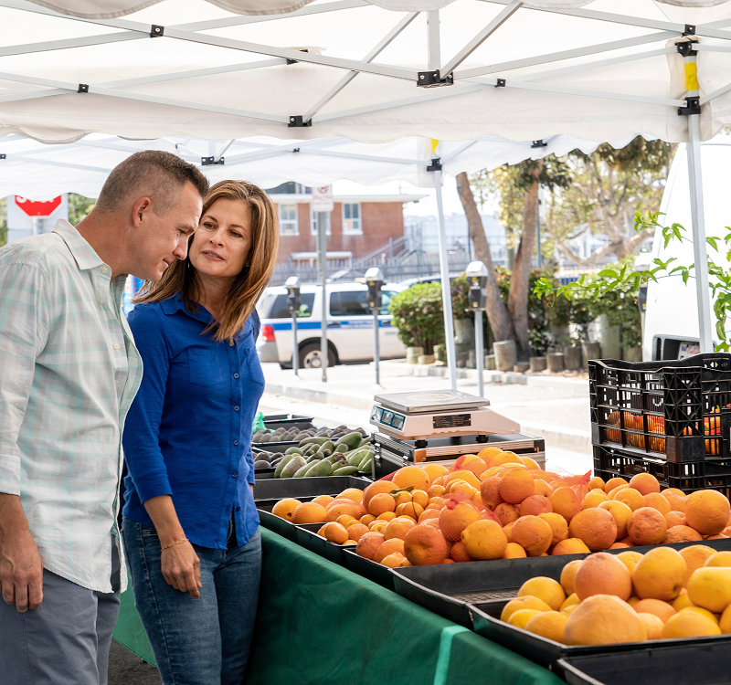 A man and woman stand beside a table filled with fresh oranges, smiling and engaging in conversation.