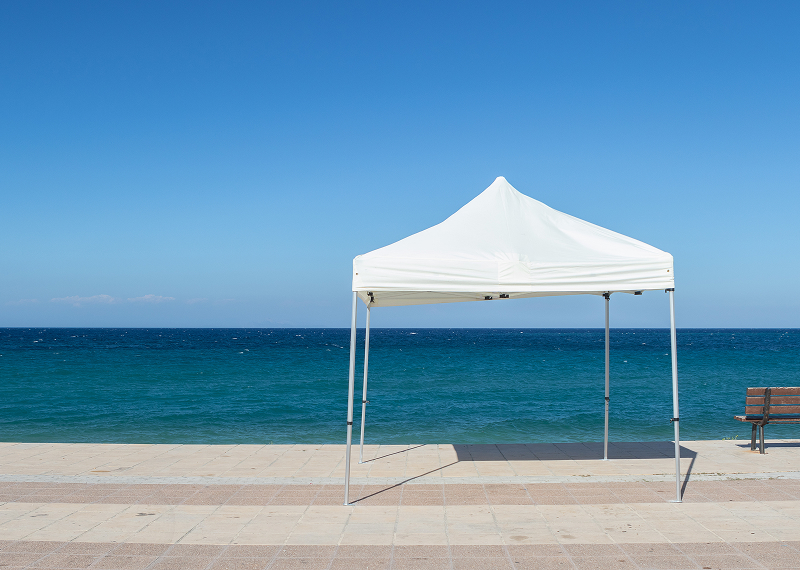 A white tent on a sandy beach, accompanied by a wooden bench nearby, with gentle waves in the background.