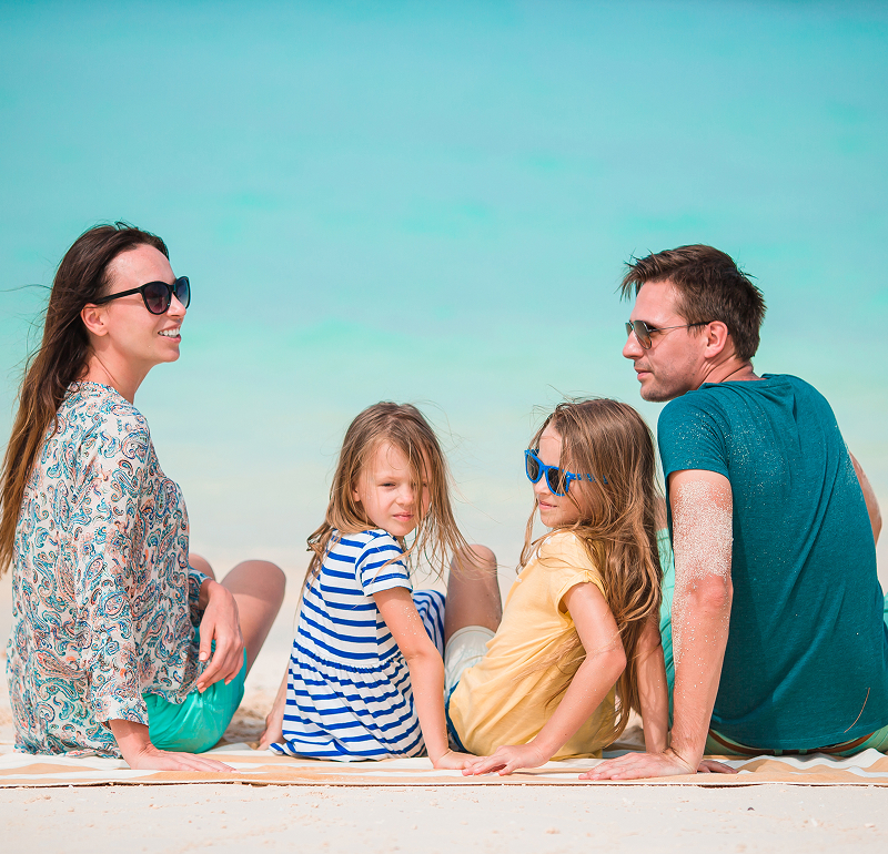 A family of four sitting on a sandy beach, enjoying the sun, with a young child playing beside them.