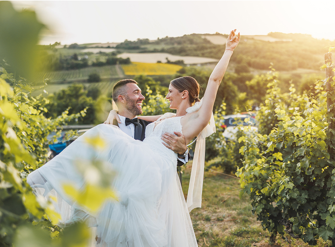 A bride and groom embrace in a vineyard during sunset, surrounded by lush vines and a warm, golden sky.