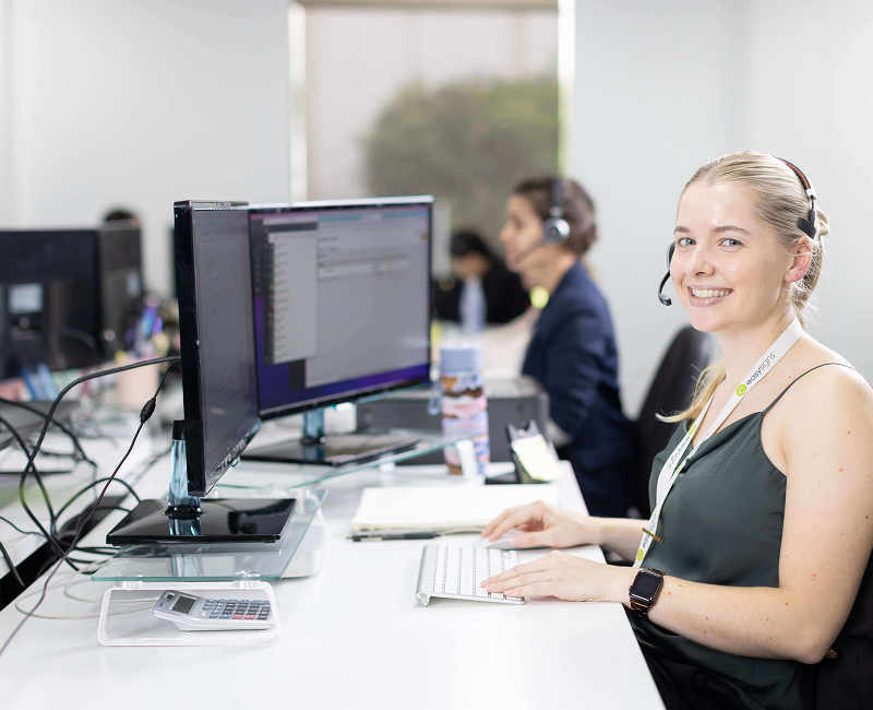 A woman seated at a desk, working on two computers, focused on her tasks in a well-lit office environment.