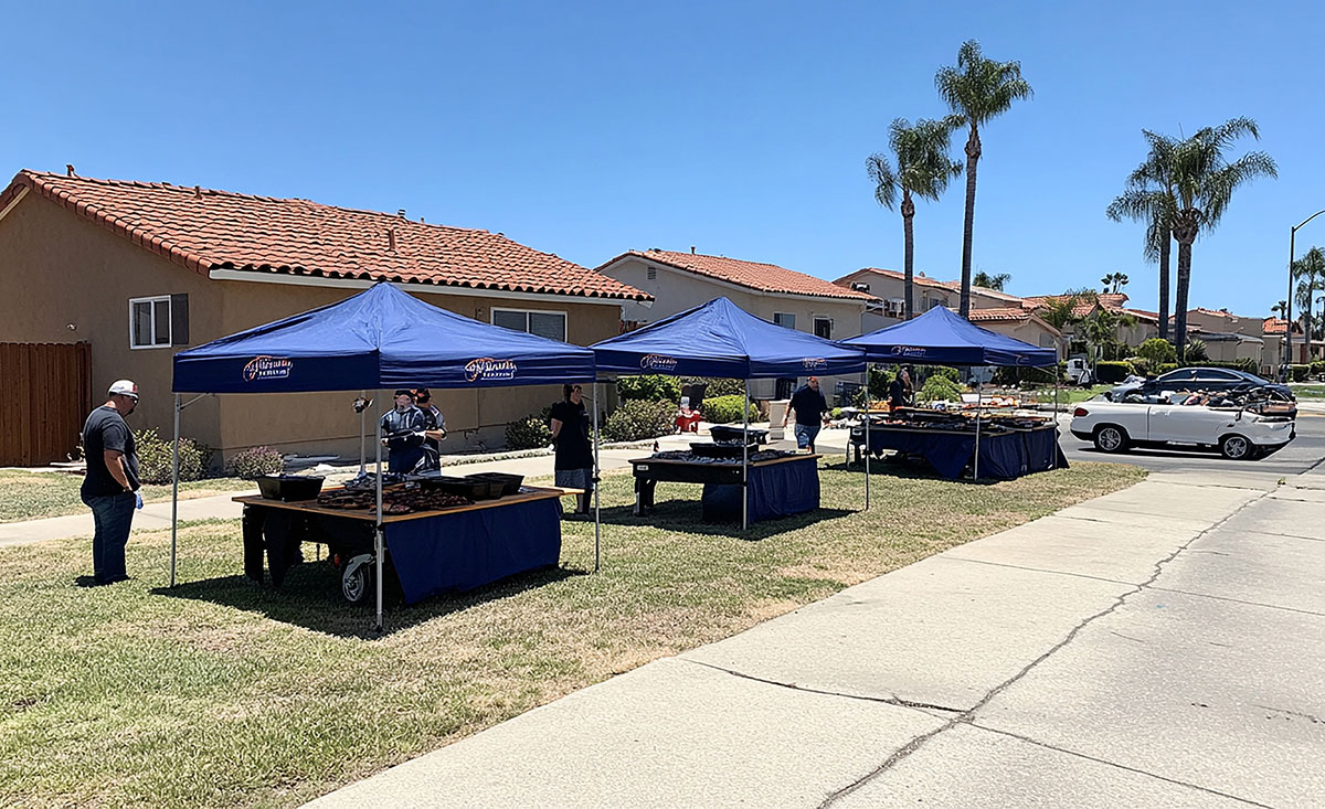 People seated at tables under blue tents, engaged in conversation and enjoying a social gathering outdoors.